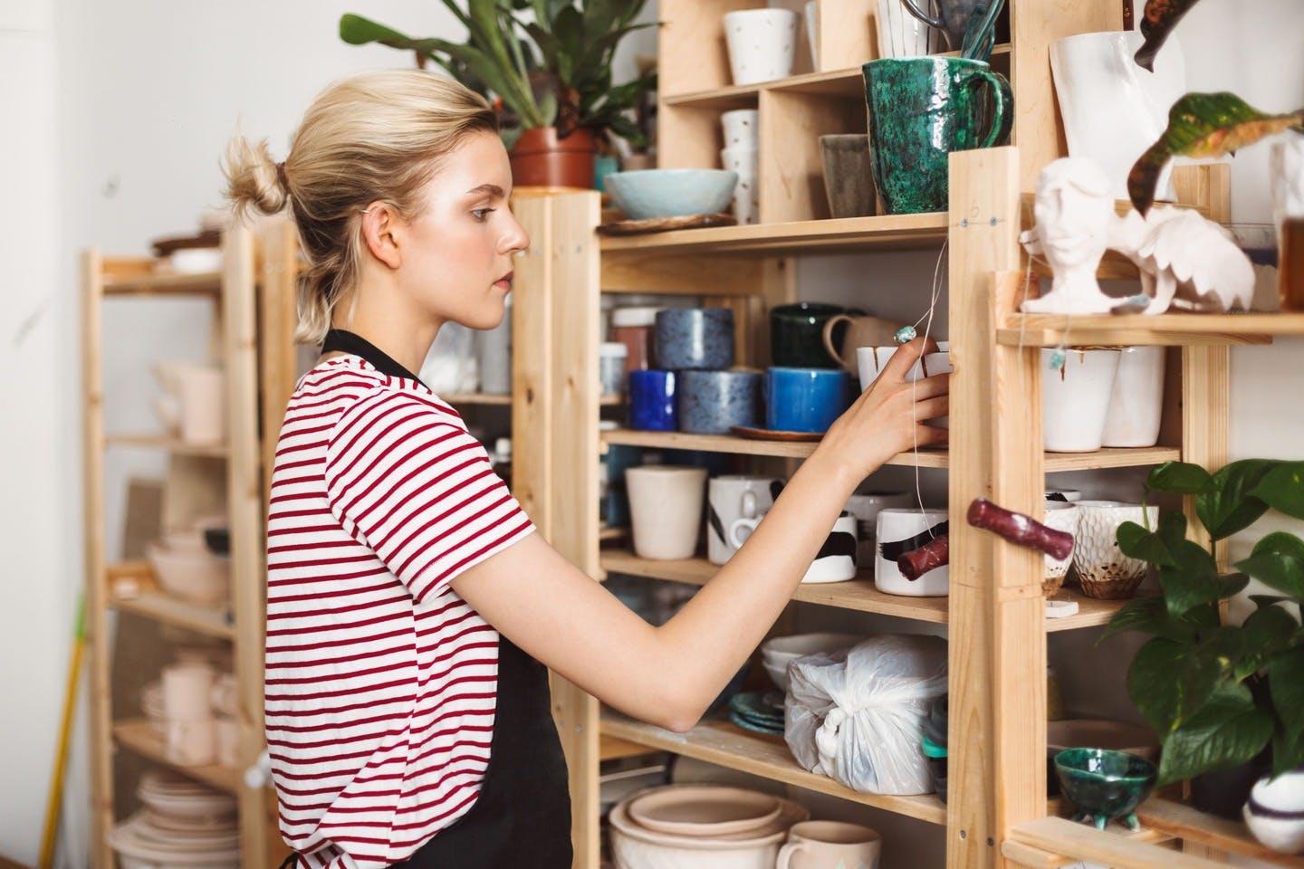 beautiful-girl-black-apron-striped-t-shirt-thoughtfully-putting-handmade-dishes-shelves-modern-pottery-studio.jpg